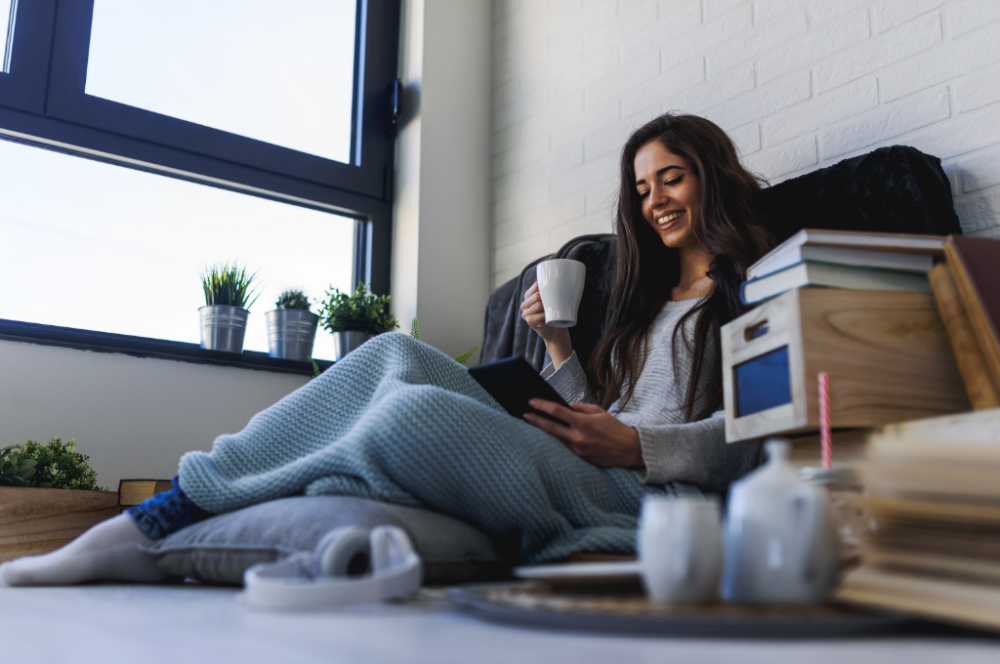A woman with long brown hair leans against a wall. She has a blanket over her legs and has a mug of coffee in one hand and a device in the other. She is smiling as she reads her ebook.