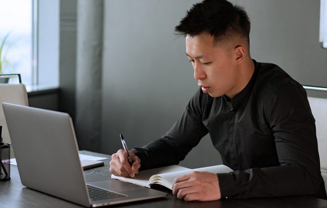 man with short black hair sitting at a desk in front of a laptop with a notebook and pen