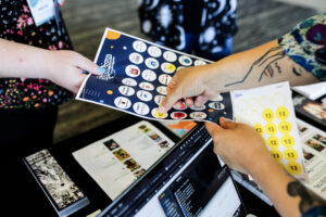 A person hands a passport with exhibitor logos over a table. Another person "stamps" the passport