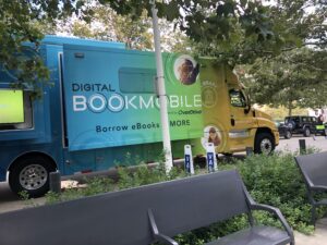 A blue, green, and yellow tractor trailer bus is parked outside. The Digital Bookmobile is a community outreach vehicle is for public libraries and schools to promote their digital catalog of ebooks, audiobooks, magazines and streaming video to their communities