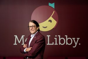 Founder and Chairman of the Board Steve Potash stands in front of burgundy wall with Libby mascot and words "Meet Libby"